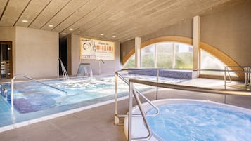 An indoor swimming pool area with two hot tubs, clear water, and a sign advertising 'Aguas de Rocallaura'.