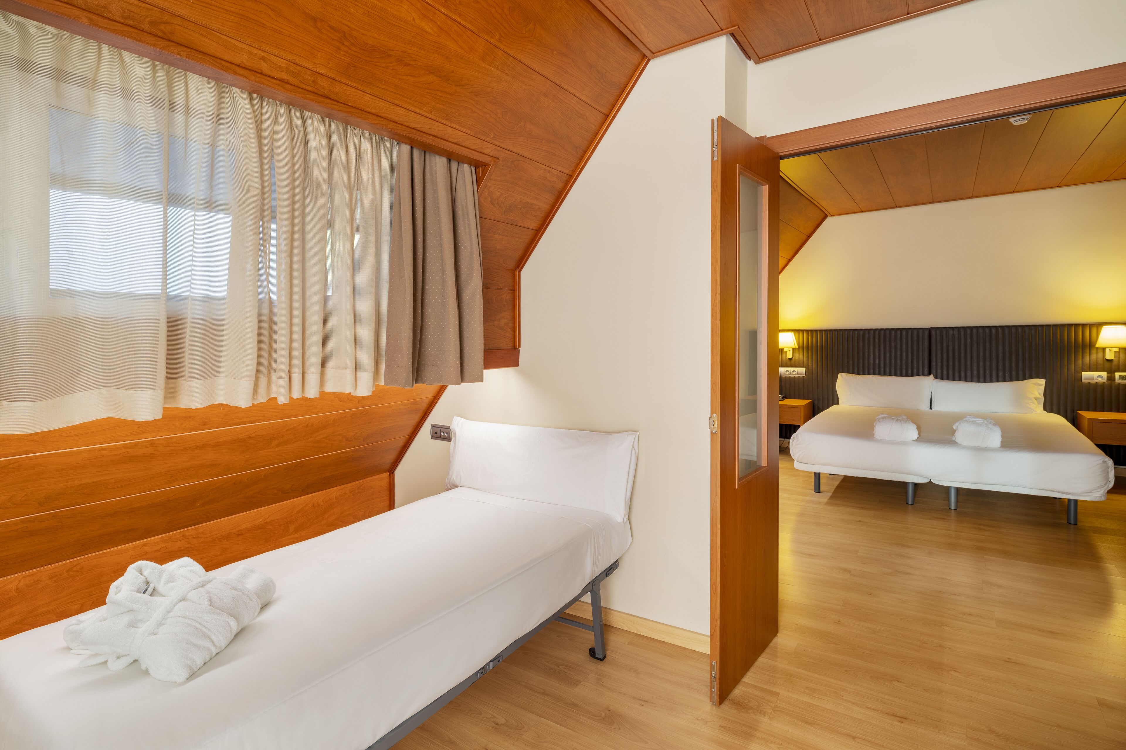 A single-bed room with wooden paneling, a window with sheer curtains, and a view into an adjacent room with a bed and nightstands.
