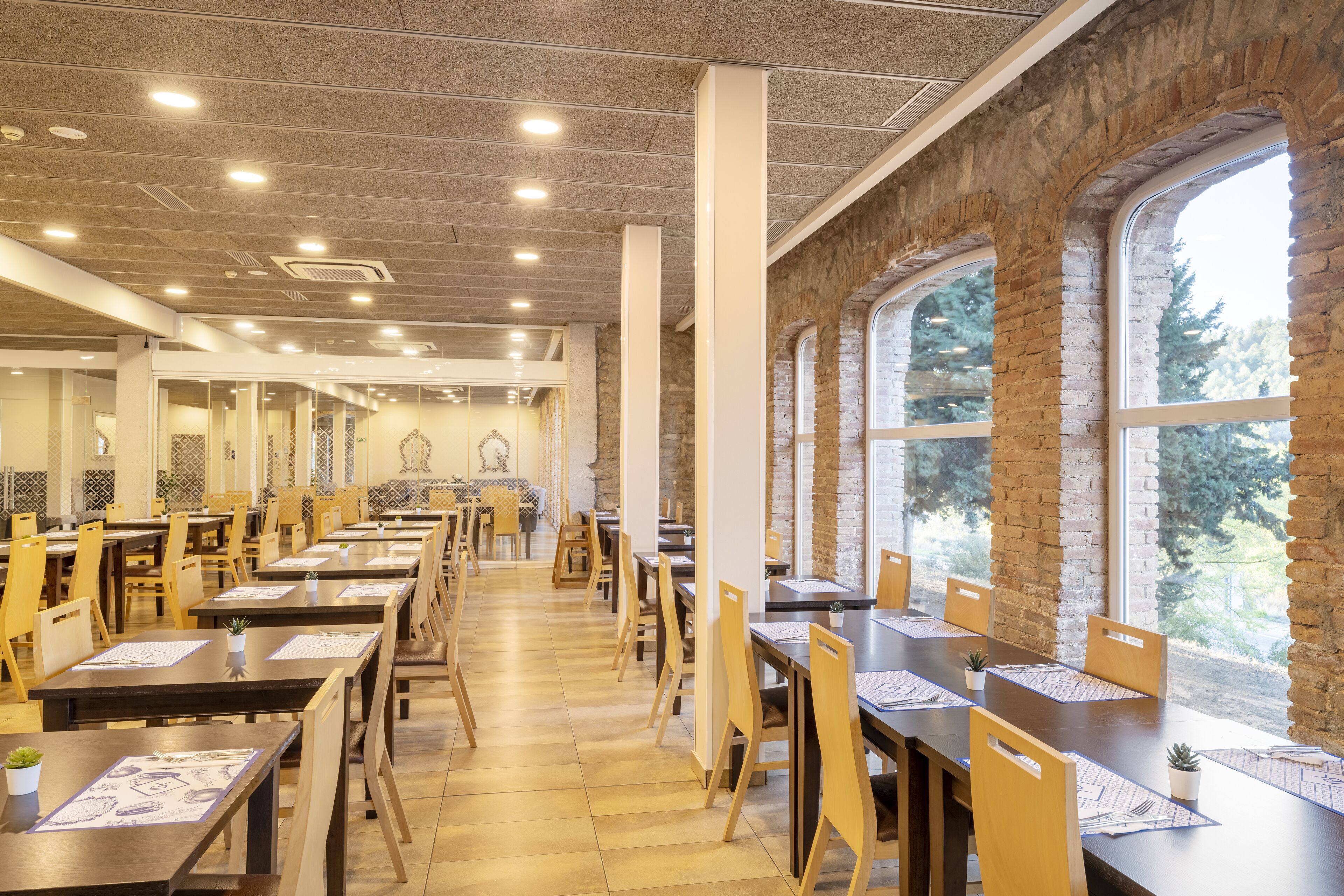 A spacious dining area with high tables and chairs, large windows with a view of greenery, and a modern ceiling with recessed lighting.