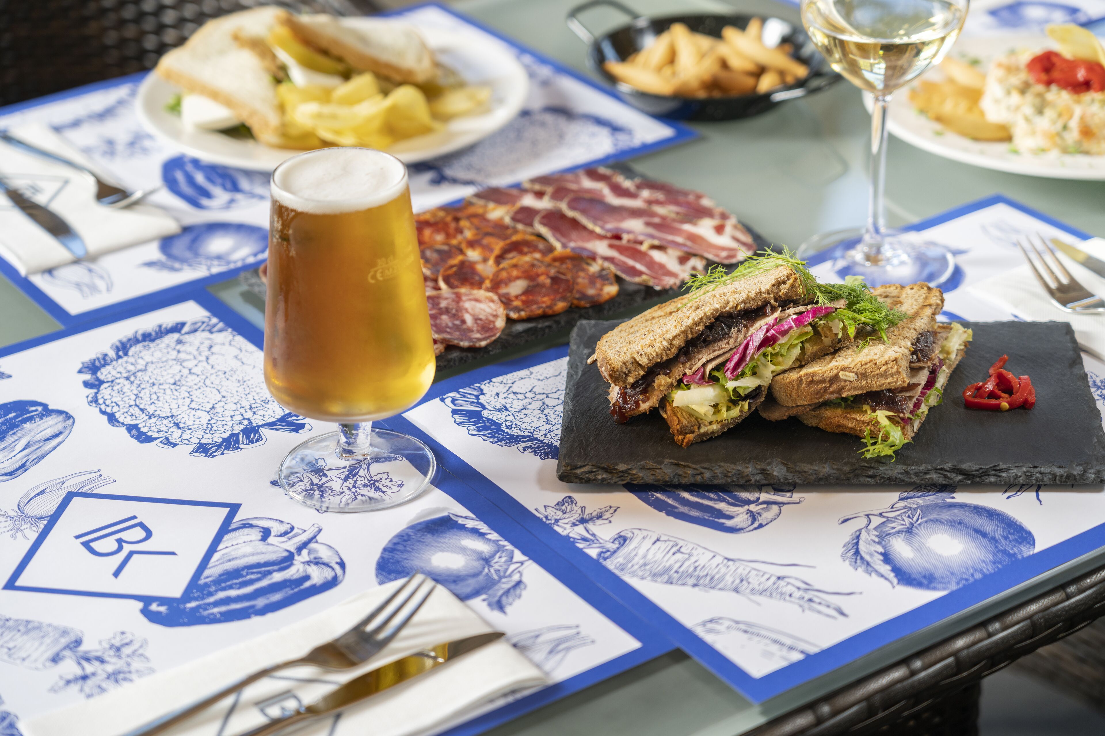 Et bord dækket med forskellige retter, herunder sandwiches, pommes frites og et glas øl, på et bord med et blå og hvidt ternet dug.