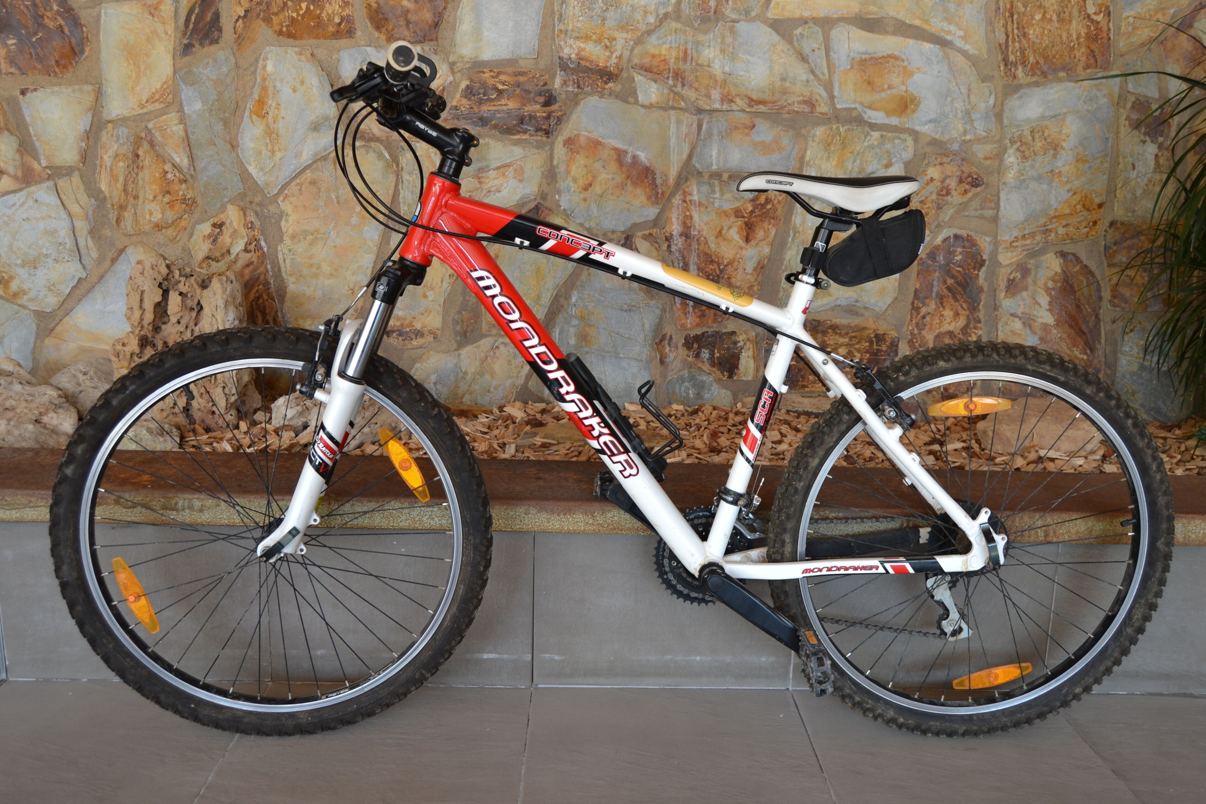 Una bicicleta de montaña con estructura blanca y detalles rojos, estacionada frente a un muro de piedra.