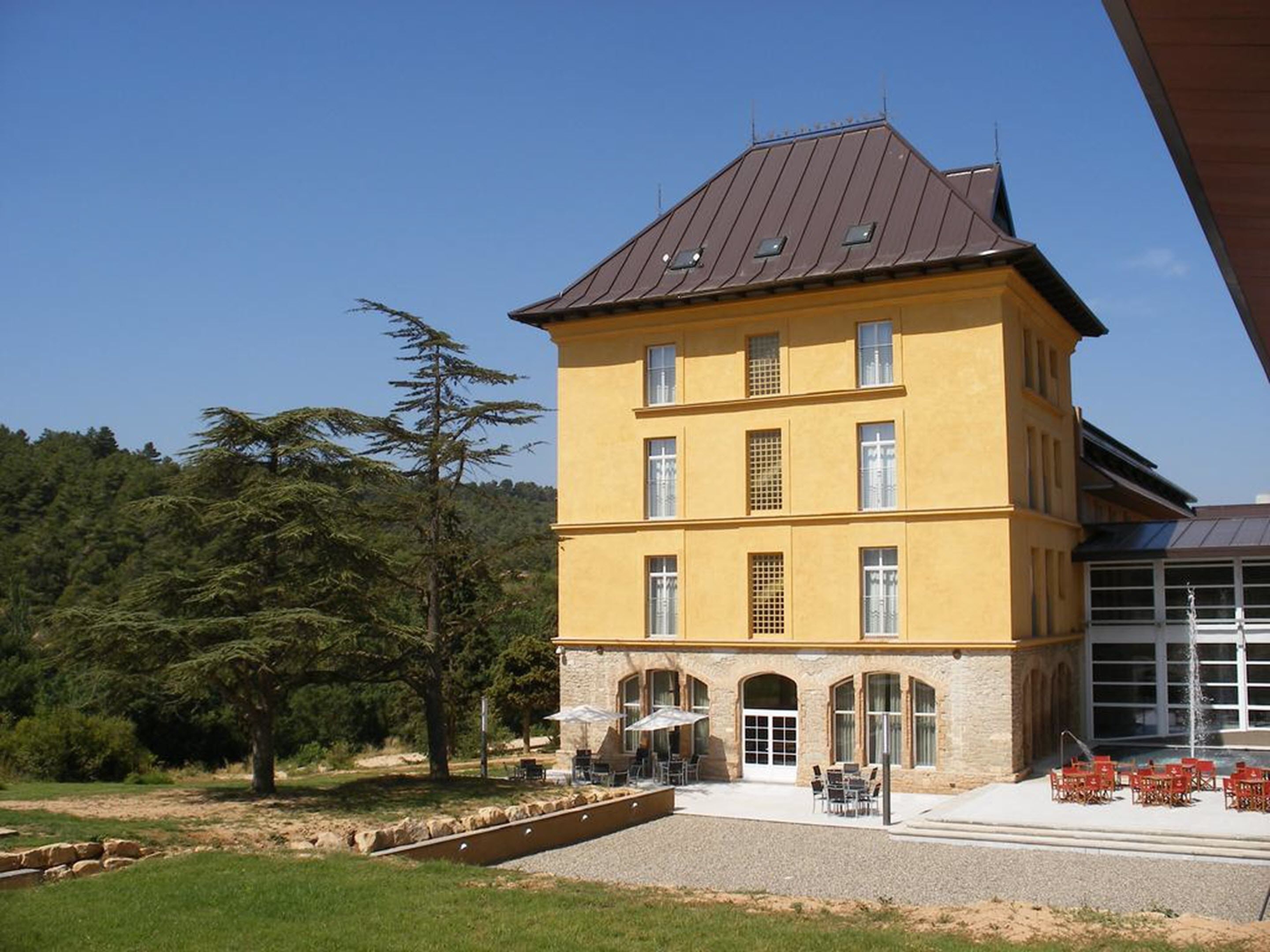 A multi-story building with a yellow facade and a brown roof, featuring outdoor seating and a small pool area.