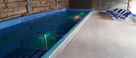 An indoor swimming pool with a stone wall, a tiled floor, and blue lounge chairs along the side.