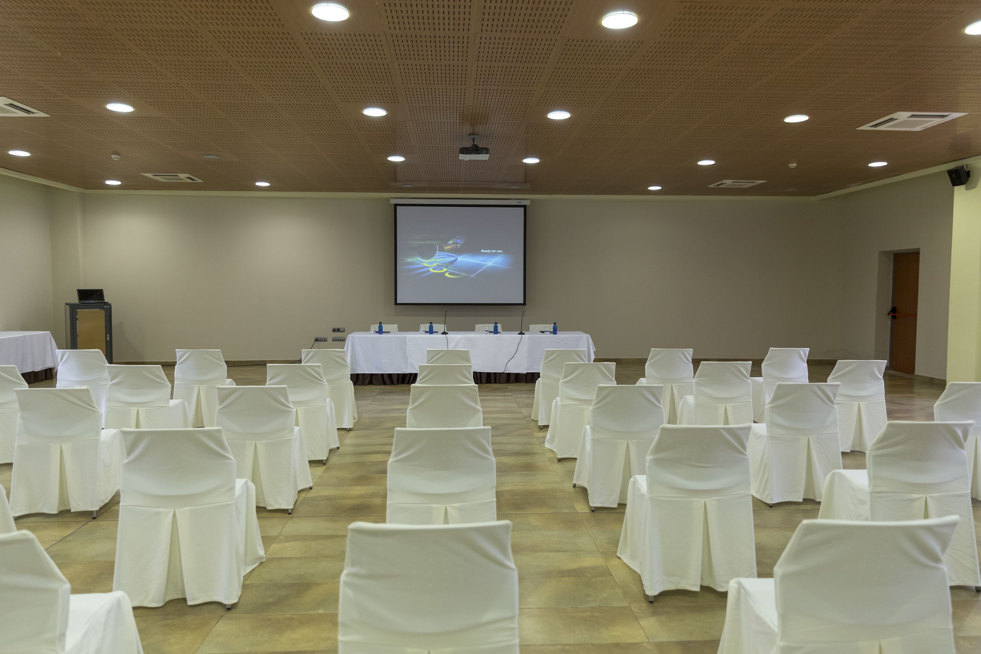 Una sala conferenze con file di sedie bianche, uno schermo per proiettore e tavoli coperti da tovaglie bianche.
