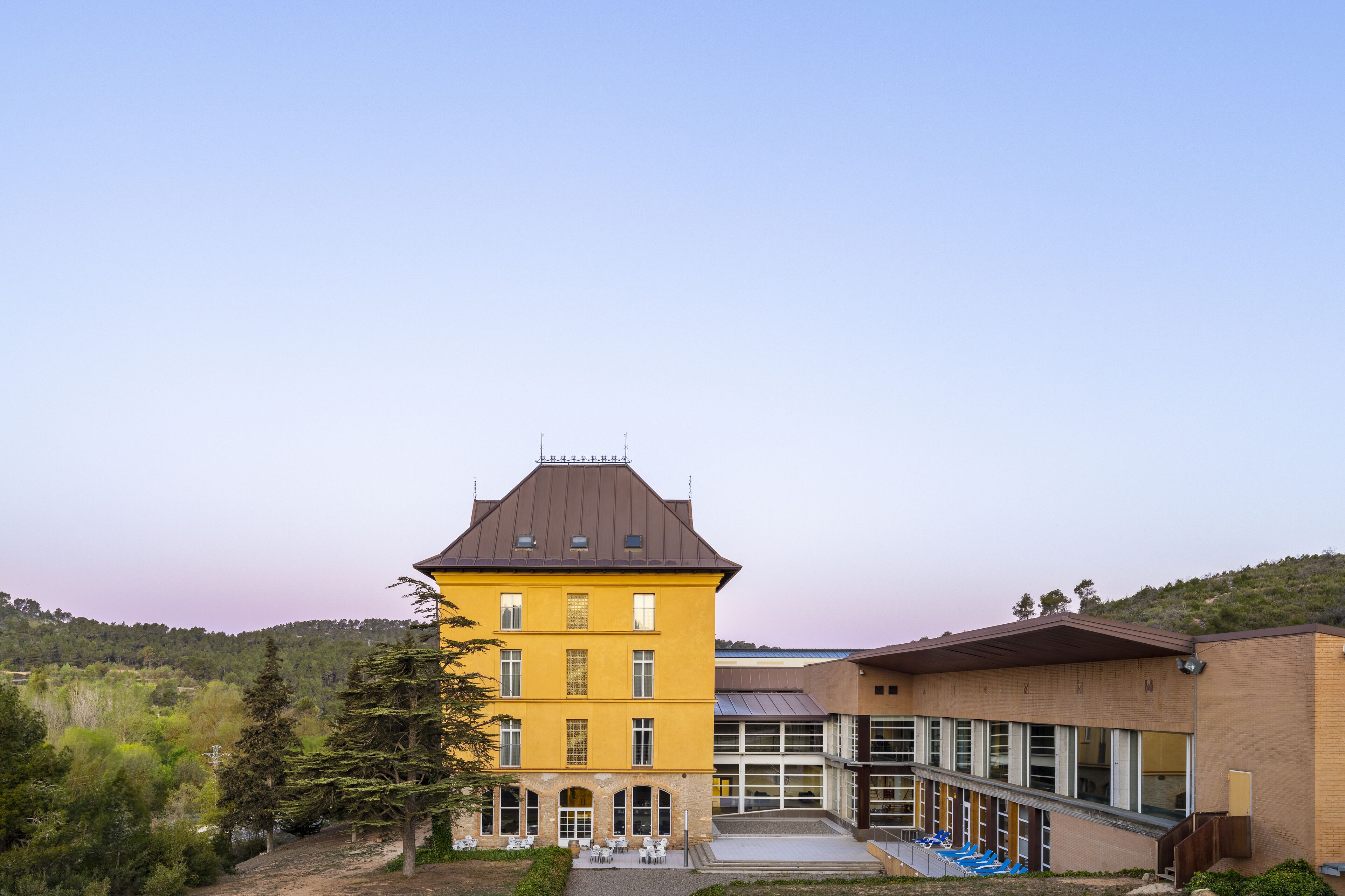 A yellow building with a brown roof, a courtyard with outdoor seating, and a view of a hilly landscape.