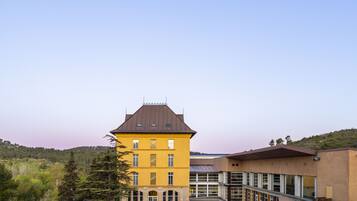 A yellow building with a brown roof, a courtyard with outdoor seating, and a view of a hilly landscape.