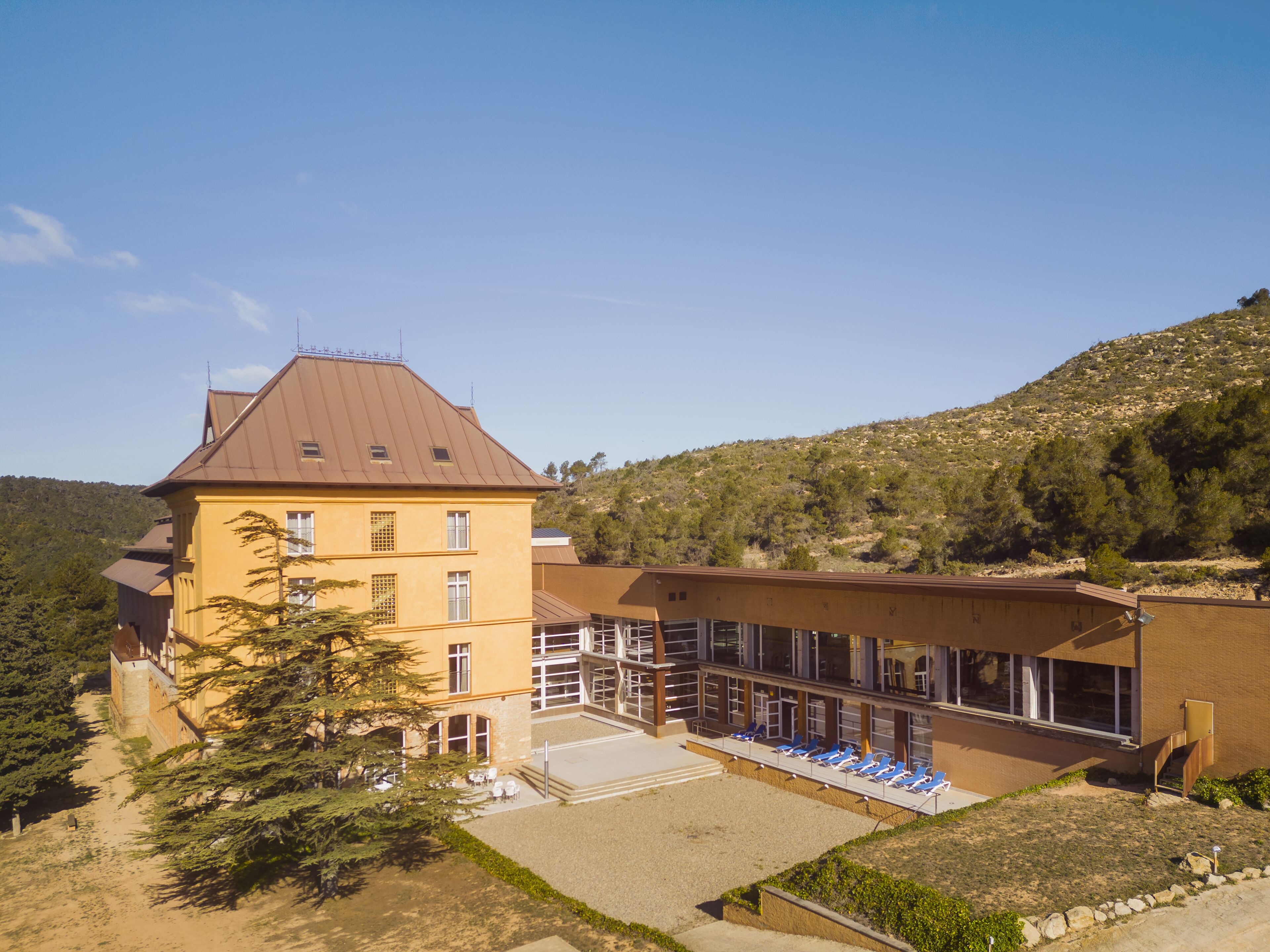 A large yellow building with multiple windows and a brown roof, surrounded by trees and a hilly landscape.