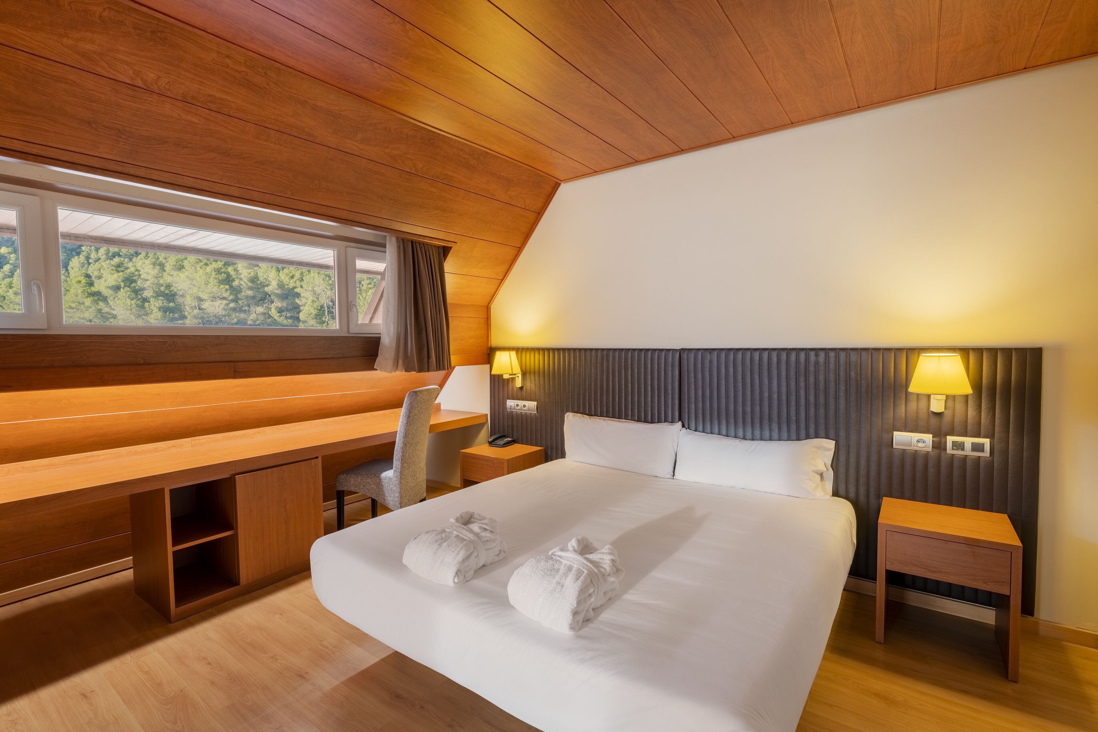 A hotel room with a large bed, a wooden desk, a chair, a bedside table, and a nightstand.