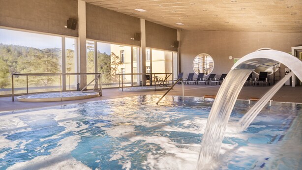 Une piscine intérieure agrémentée d’une cascade, de grandes fenêtres offrant une vue sur l’extérieur et un espace détente avec des fauteuils.