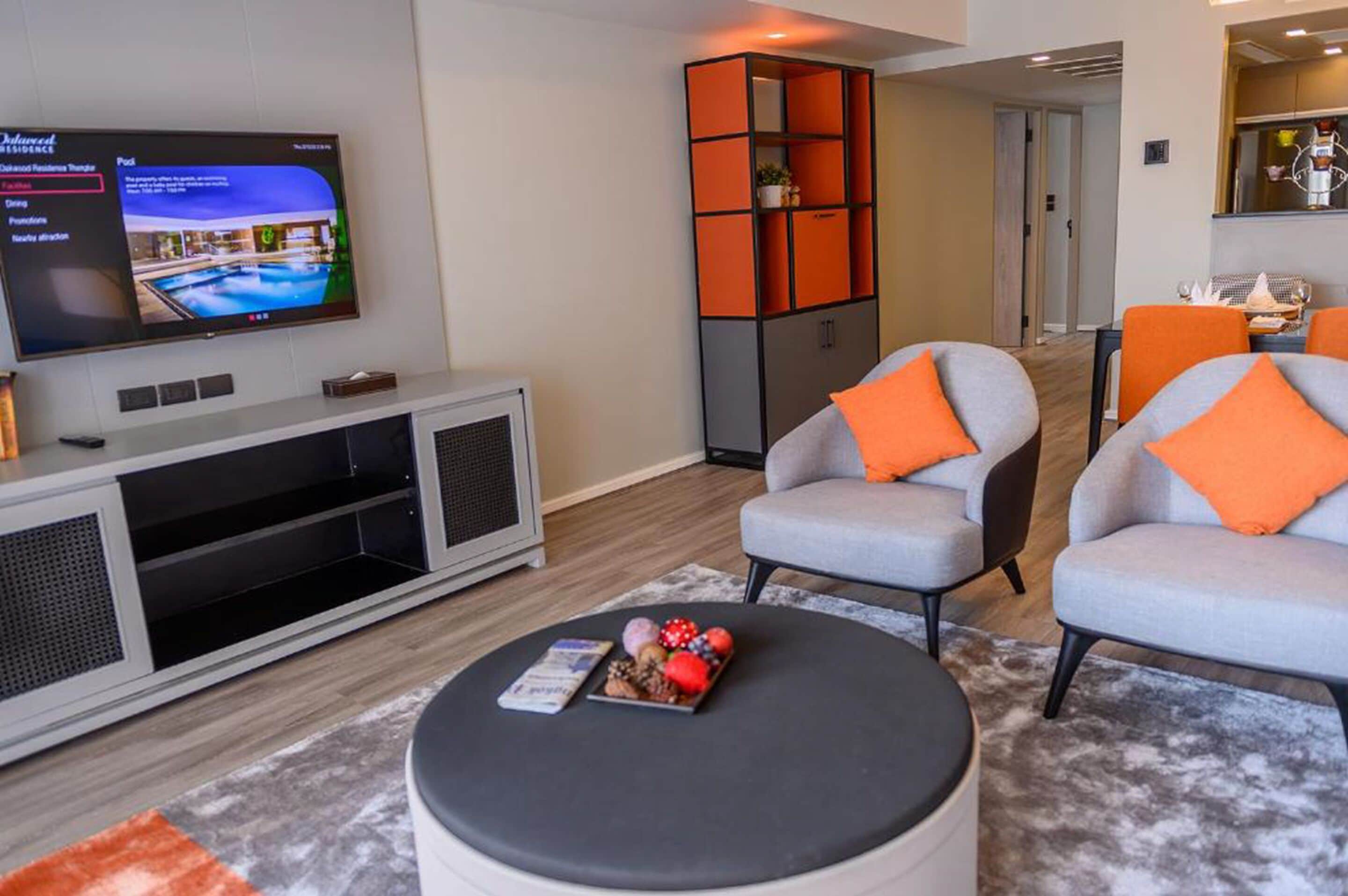 A modern living room with a flat-screen TV, grey armchairs with orange cushions, a black ottoman, and a grey sofa.