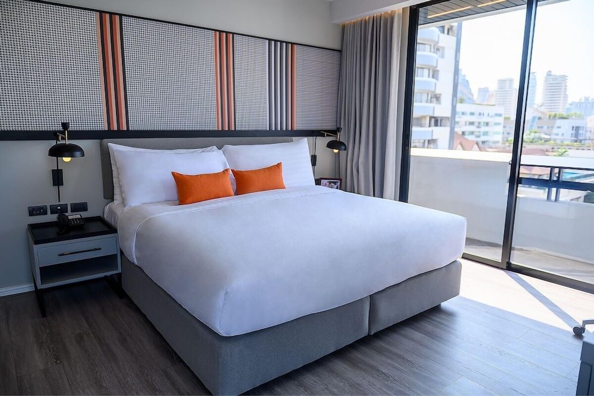 A modern hotel room with a large bed, a nightstand, a wall-mounted lamp, and a view of the city through large windows.