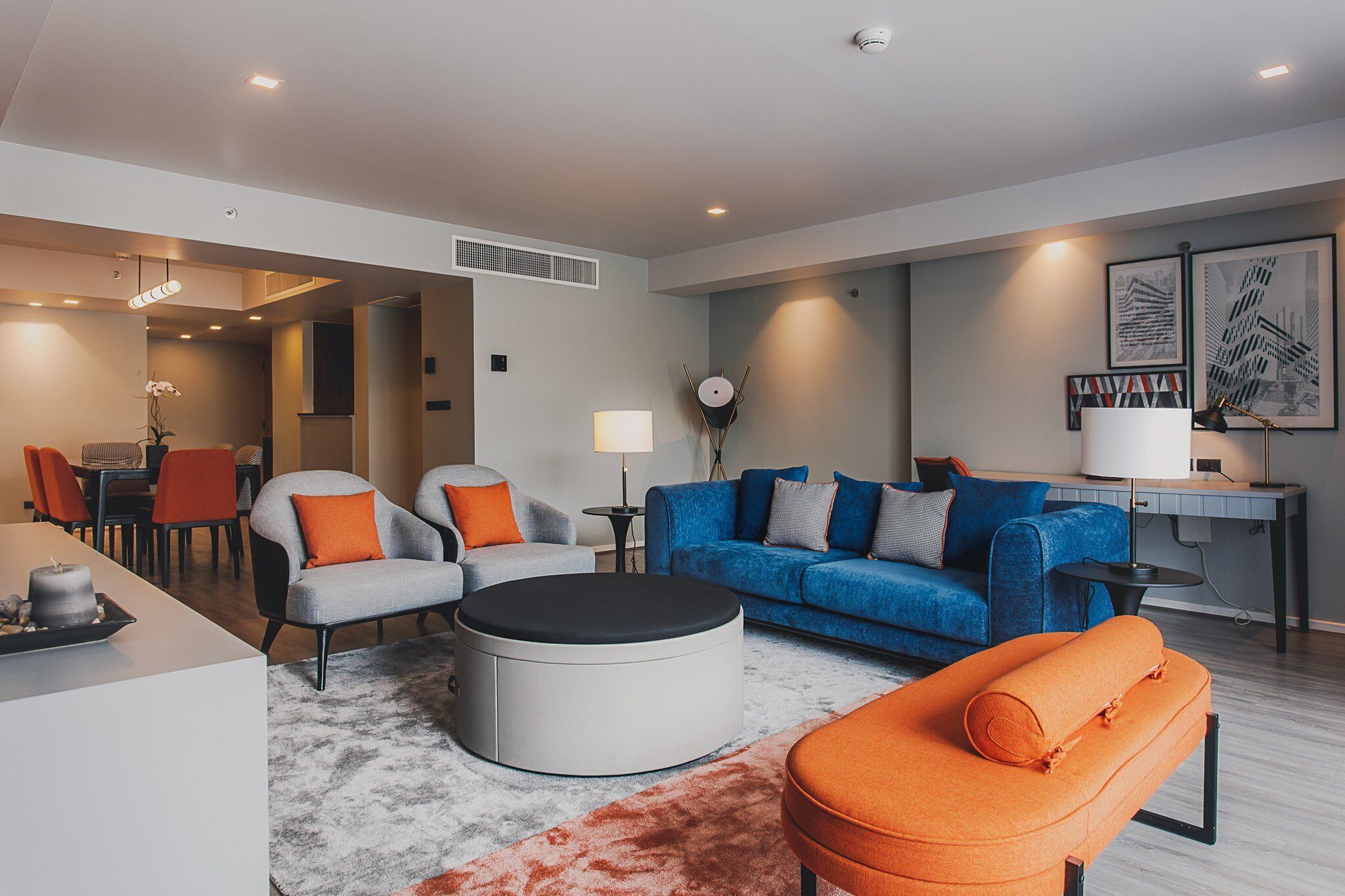 A modern living room with a blue sofa, grey armchairs, and an orange ottoman. There is a round coffee table, a dining area with chairs, and a desk with a lamp.