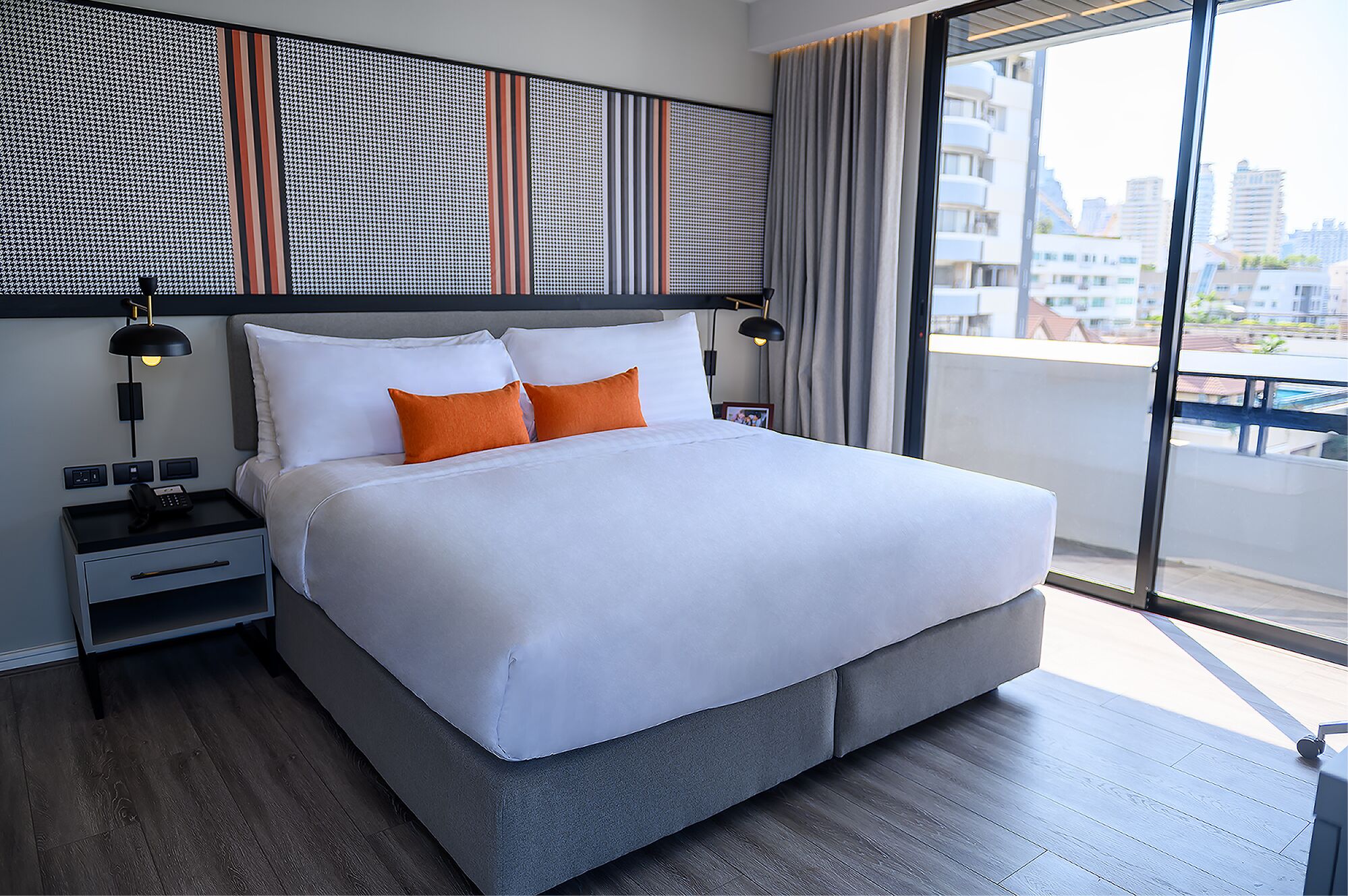 A modern hotel room with a large bed, two bedside tables, a phone, and a view of the city through large windows.