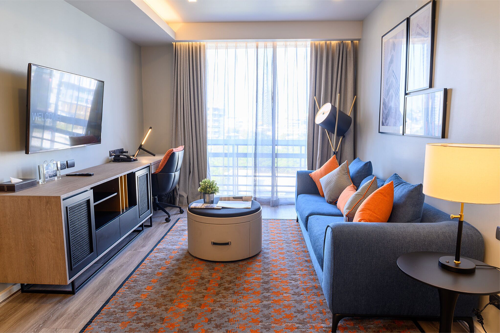 Una habitación de hotel moderna con una televisión de pantalla plana, un sofá con cojines naranja, un otoman redonda y una mesita de noche con una lámpara.