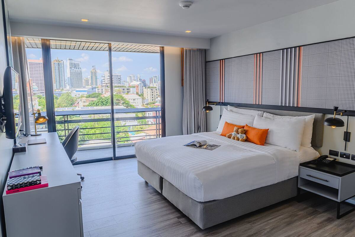 Une chambre d’hôtel moderne dotée d’un grand lit, d’un bureau et offrant une vue sur les toits de la ville grâce à de grandes fenêtres du sol au plafond.