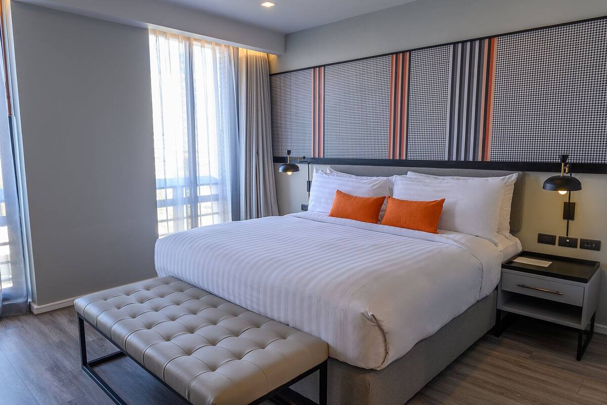 Une chambre d’hôtel moderne avec un grand lit, une banquette, une table de chevet et une tête de lit décorative.
