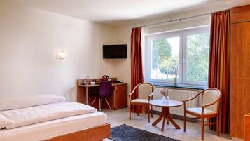 A hotel room with a bed, a desk with a chair, a small table with chairs, a TV mounted on the wall, and a window with curtains.