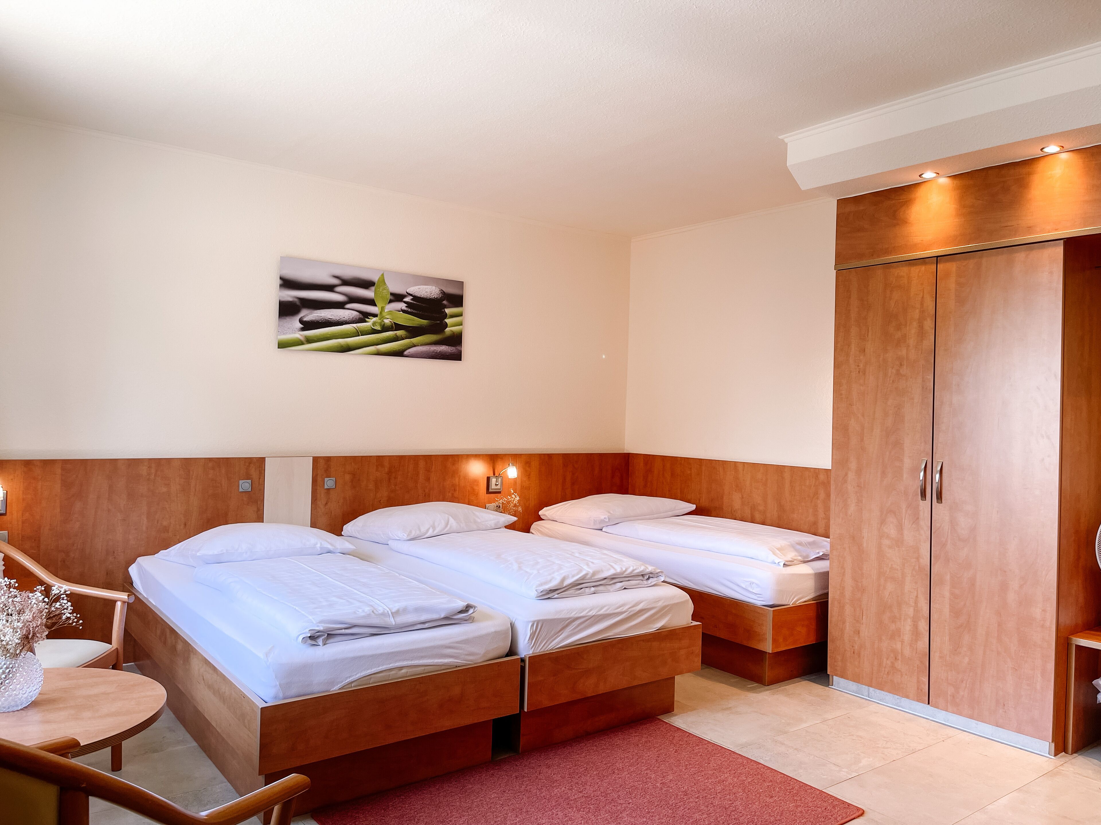 Habitación de hotel con dos camas, un armario de madera, una mesita con un florero y un cuadro colgado en la pared.