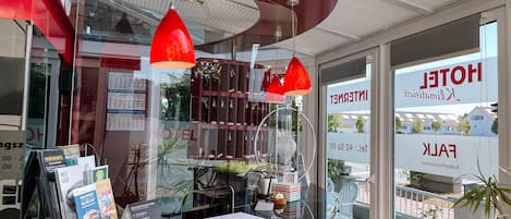 Zona de recepción de un hotel con un mostrador de cristal, luces colgantes rojas y vista al acceso del hotel.