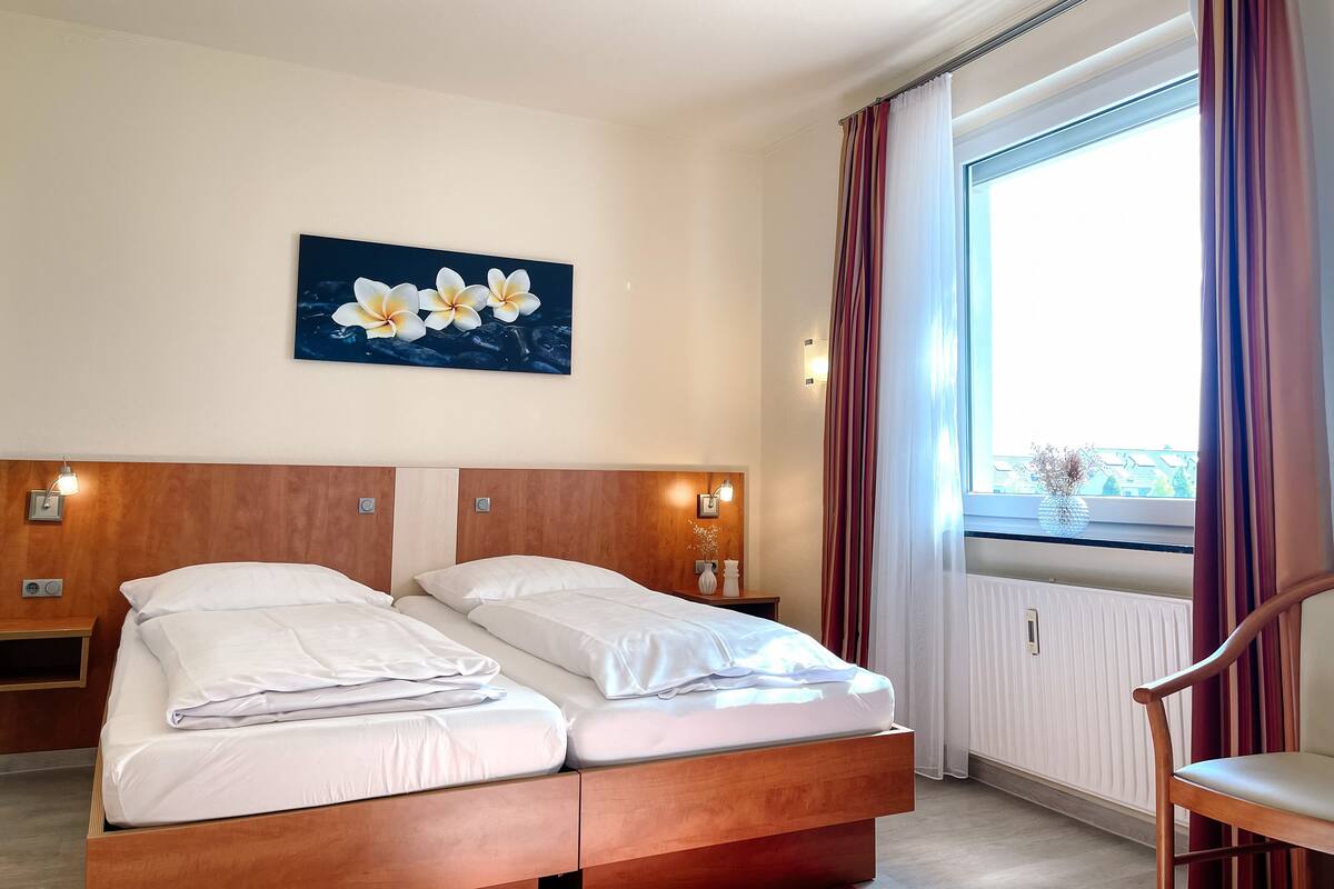Hotellihuone, jossa on puinen sänky, kaksi yöpöytää valaisimineen, ikkuna verhoineen ja seinälle ripustettu kukka-aiheinen taulu.