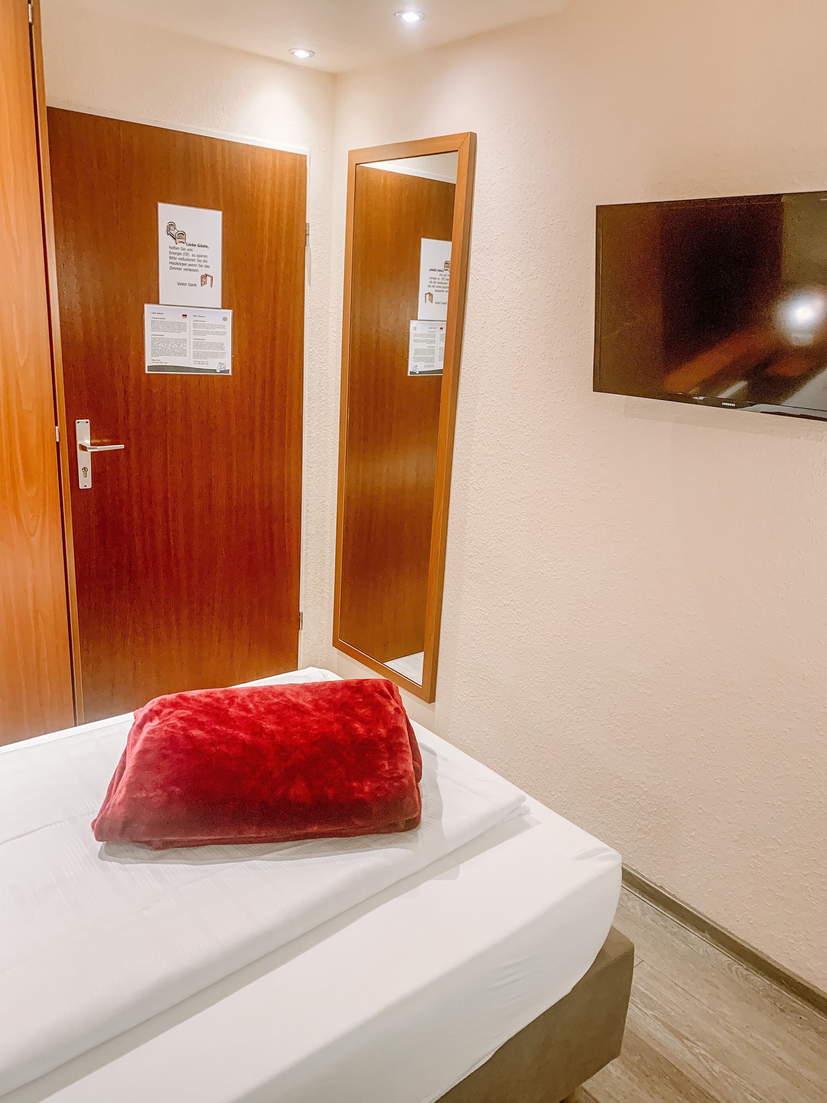 Une chambre d’hôtel avec un lit, un oreiller rouge, des portes en bois et un téléviseur fixé au mur.