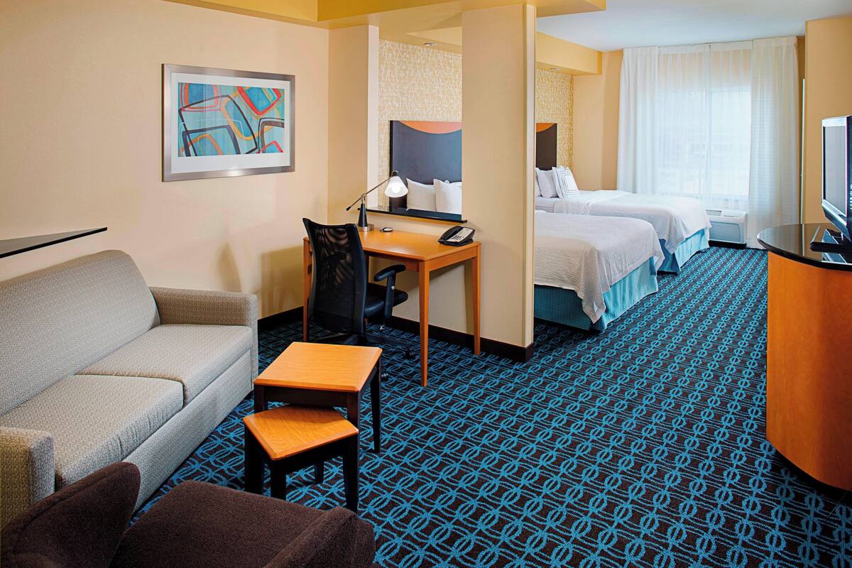 Ein Hotelzimmer mit einer Couch, einem Schreibtisch, einem Stuhl, zwei Betten, einem Fernseher und einem gemusterten Teppich.