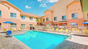 A swimming pool with lounge chairs and umbrellas, surrounded by a white fence and a building with multiple windows.