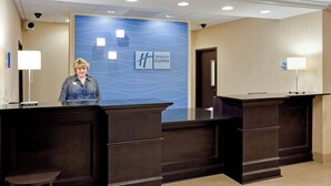 A reception desk area with a blue wall featuring a wave pattern and the Holiday Inn Express logo. There are two receptionists standing behind the desks, and a check-in counter is visible.