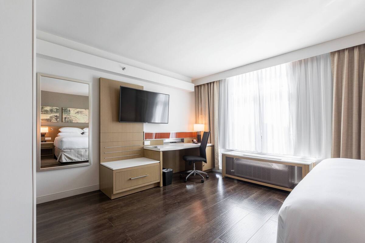 A hotel room with a bed, a desk, a chair, a TV, a window with curtains, and a radiator.