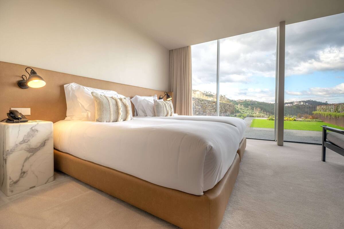 A modern hotel room with a large bed, a marble nightstand, a desk lamp, and a view of a green landscape through large windows.