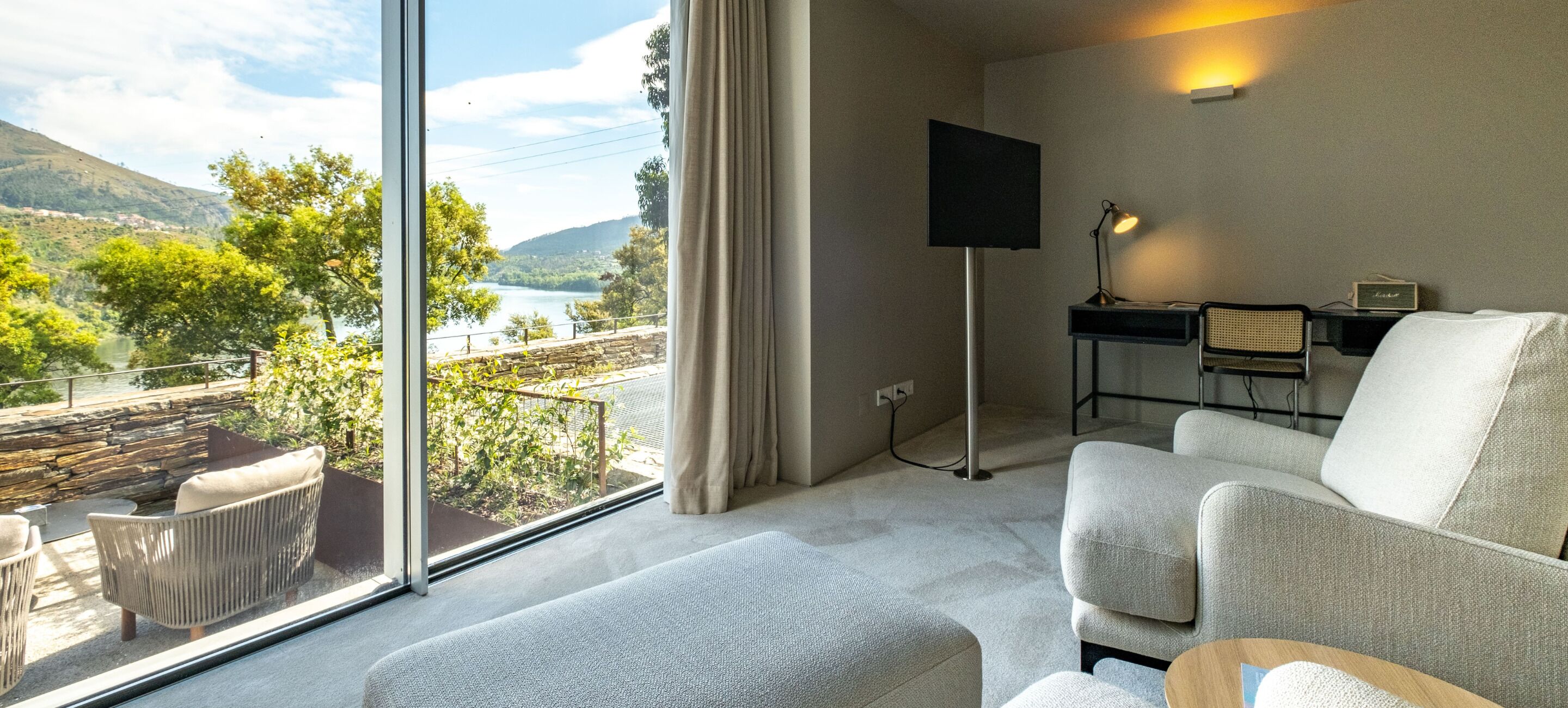 Ein modernes Hotelzimmer mit großem Fenster und einem herrlichen Blick auf einen See und Berge. Es gibt eine gemütliche Sitzecke mit einer Couch und Sesseln, einen Schreibtisch mit Stuhl sowie einen flachbildfernseher an der Wand.