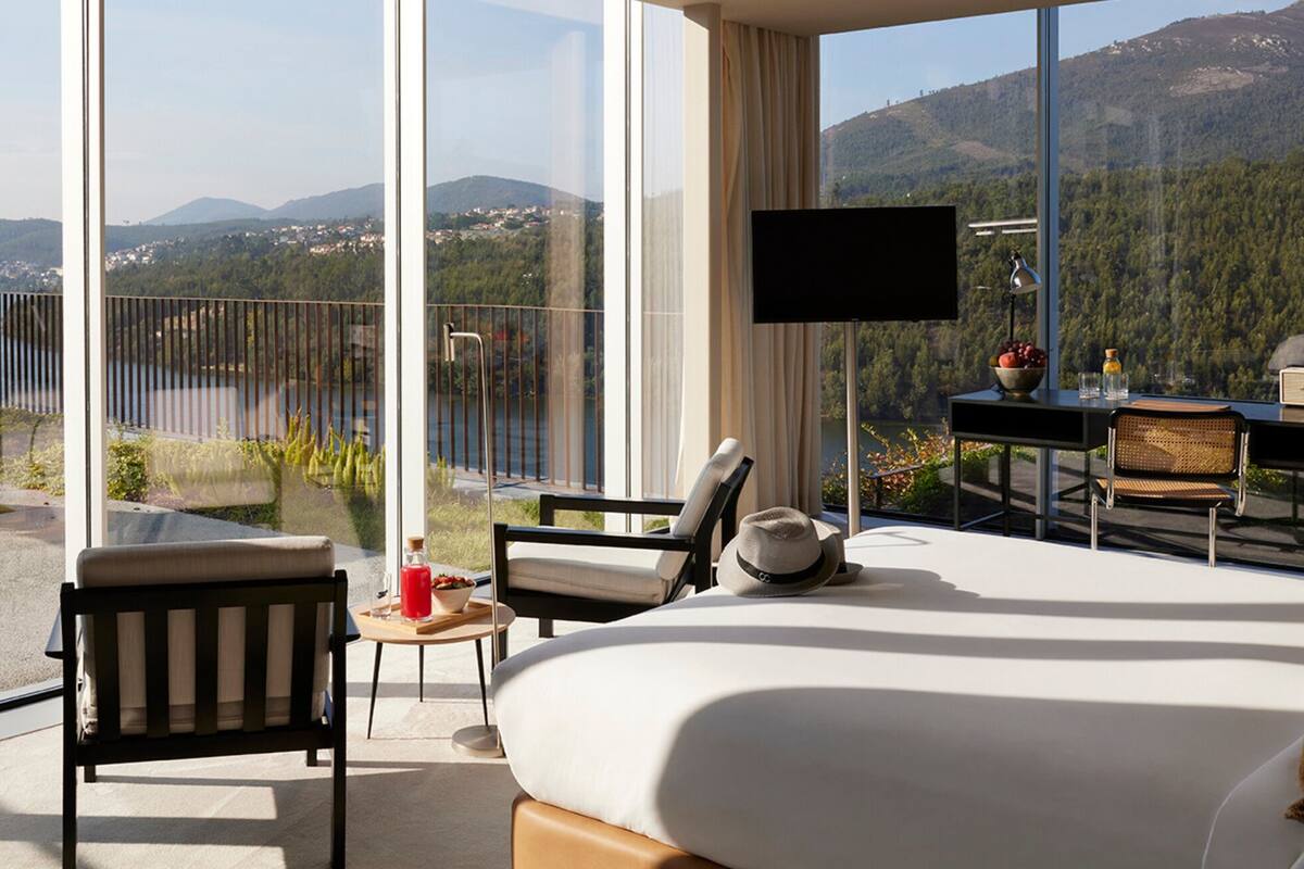 A modern hotel room with a large bed, a TV, and a balcony with a view of mountains and a lake.