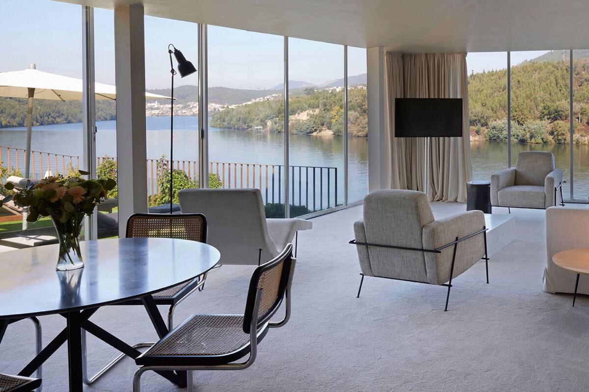A modern living room with a large glass wall, a dining table, two armchairs, a sofa, a TV, and a bathtub.