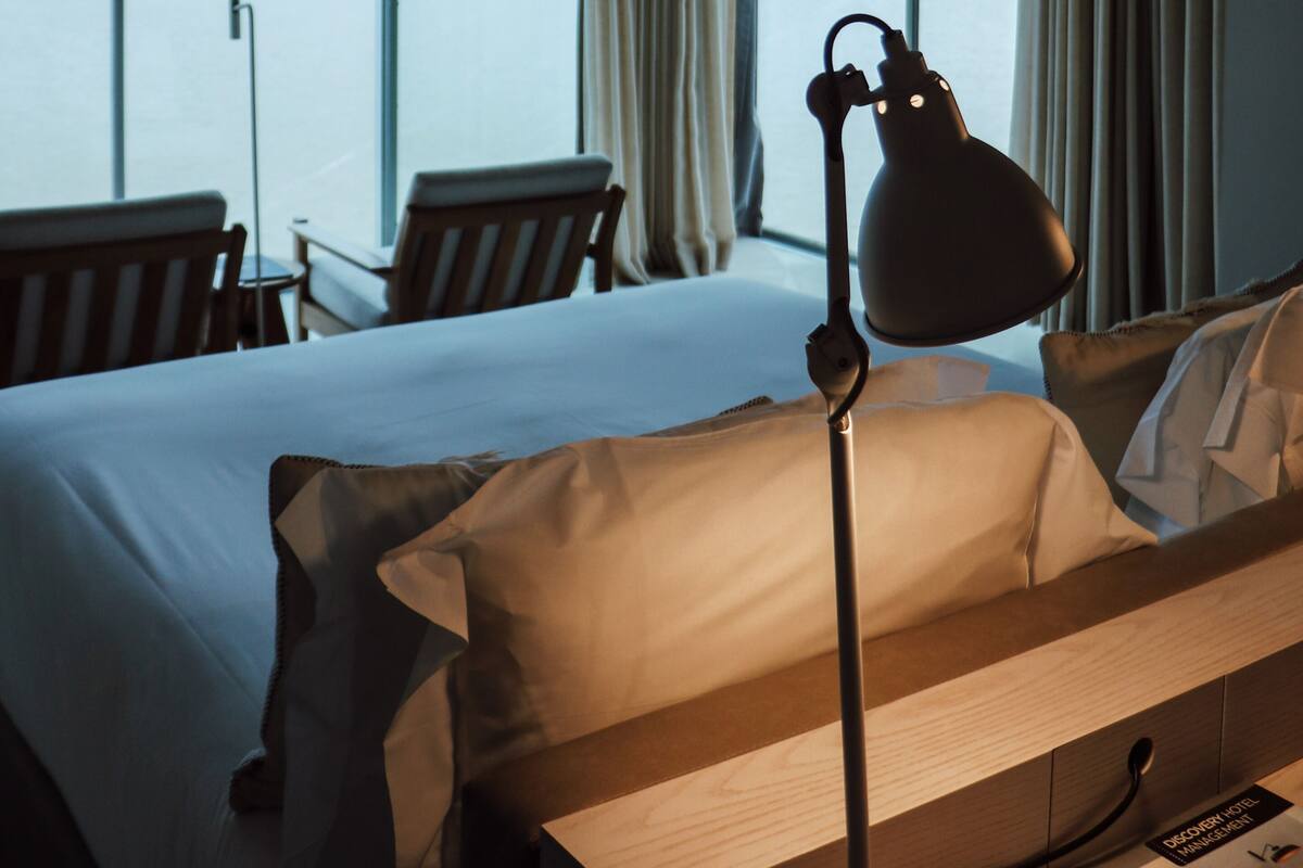 A hotel room with a large bed, a desk, a chair, a desk lamp, and a view of a lake and mountains through large windows.