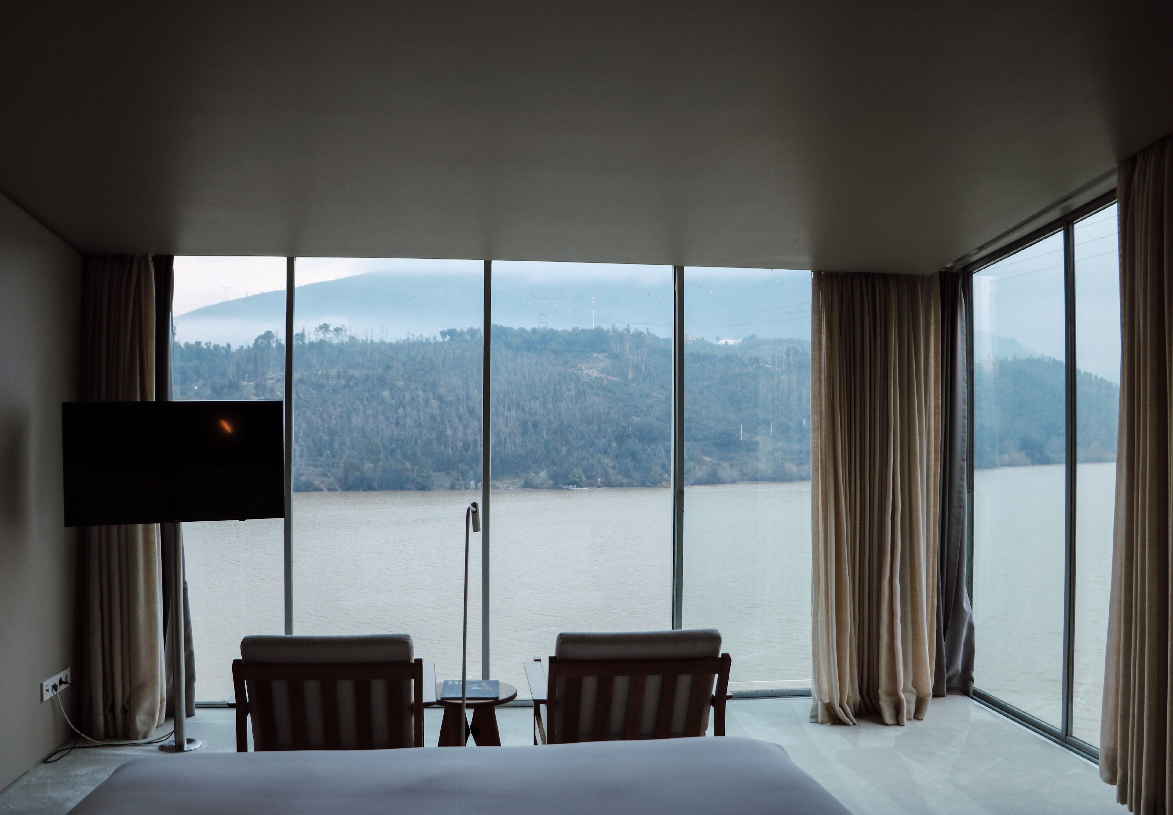 Habitación de hotel con una cama grande, dos sillas, una mesita y un televisor. La habitación tiene ventanas amplias con vistas a un lago y colinas.