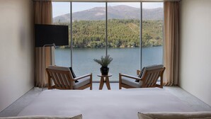 A bedroom with a large bed, two wooden chairs, a small table, and a TV. It features a large window with a view of a lake and mountains. - Octant Douro (Castelo de Paiva)