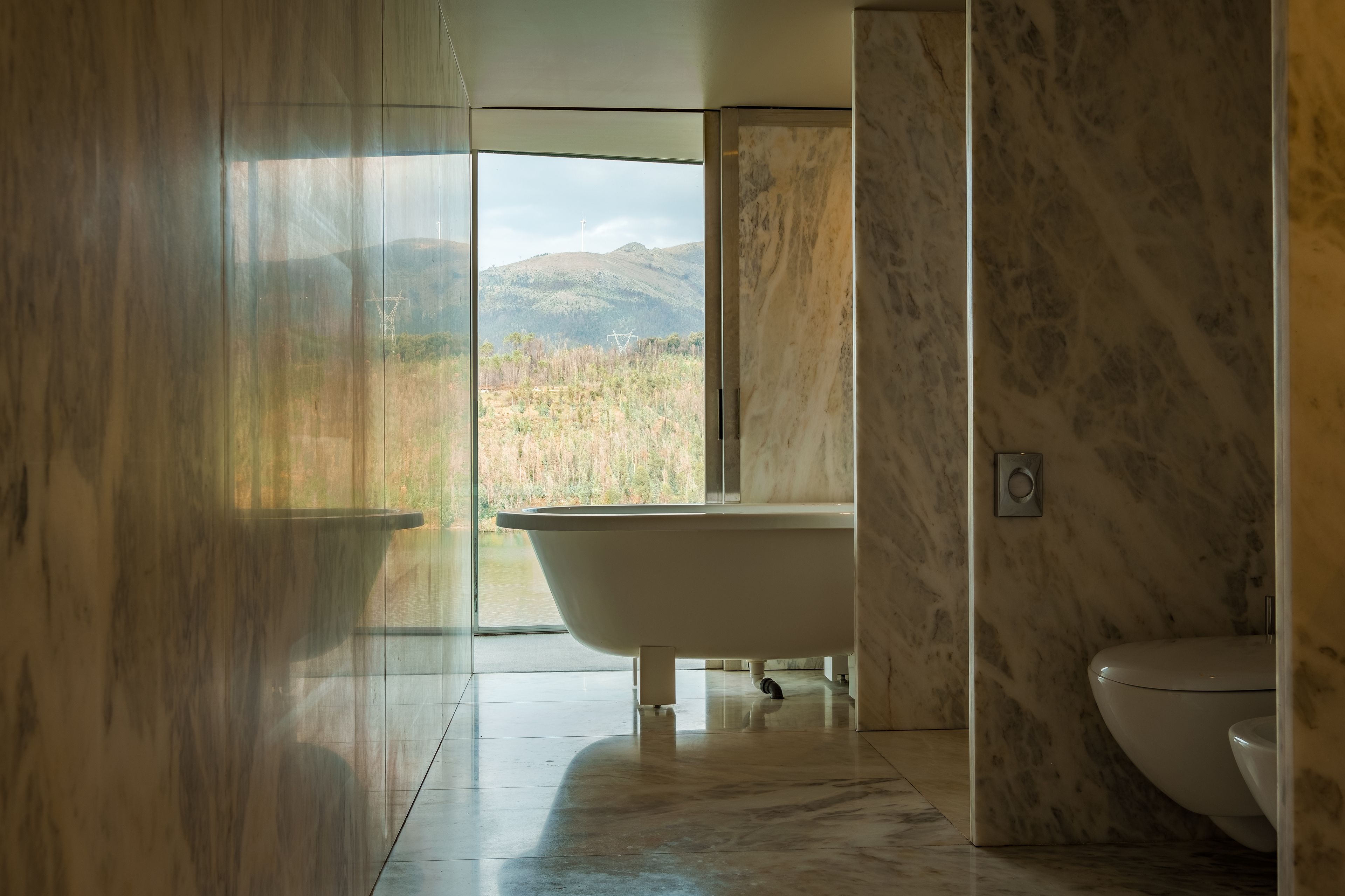 Un baño moderno con una bañera independiente, un inodoro y un amplio ventanal con vistas a un paisaje montañoso.