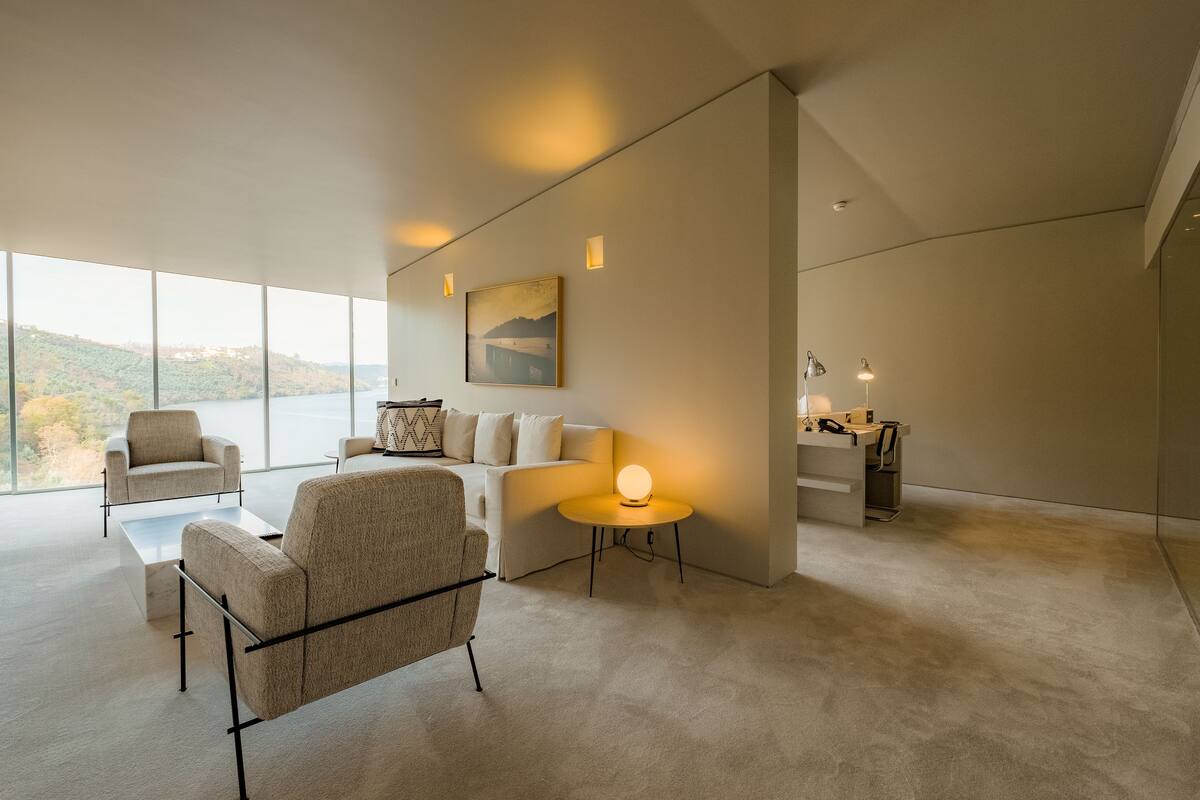 A spacious living area with a large window, a sofa, two armchairs, a coffee table, and a small side table with a lamp.