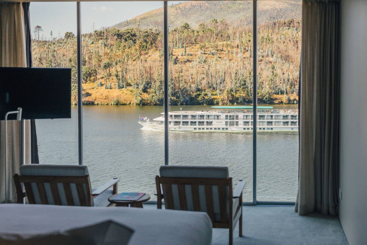 A hotel room with a large window overlooking a lake and a boat. There are two wooden chairs and a small table with a book.