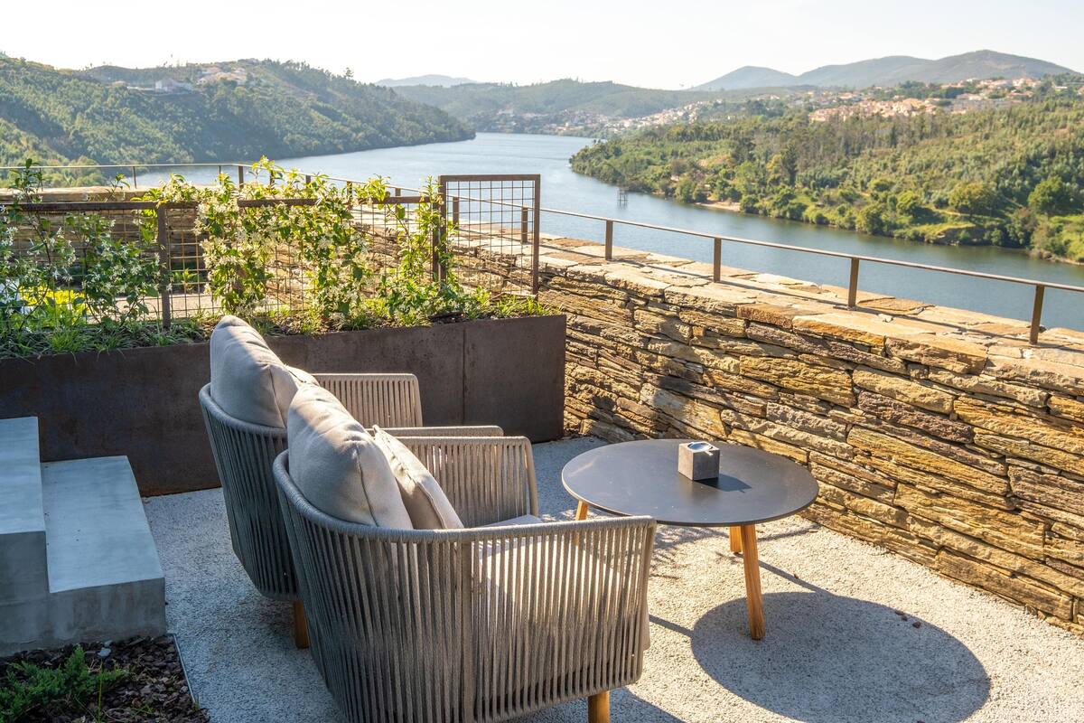 A terrace with wicker furniture and a stone wall overlooking a river.