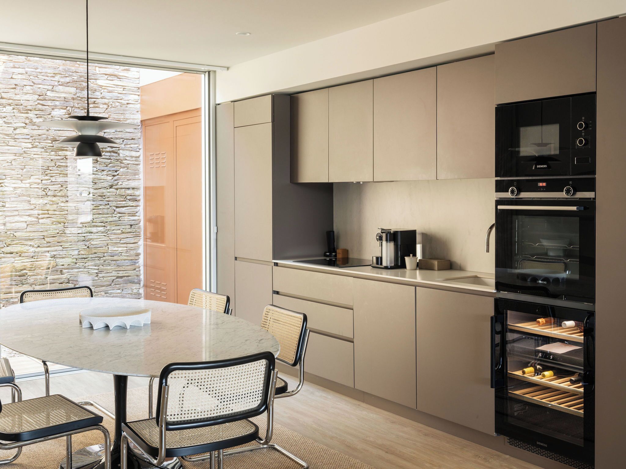 Een moderne keuken met een stenen muur, een eettafel met stoelen en inbouwapparatuur, waaronder een koffiezetapparaat en een oven.