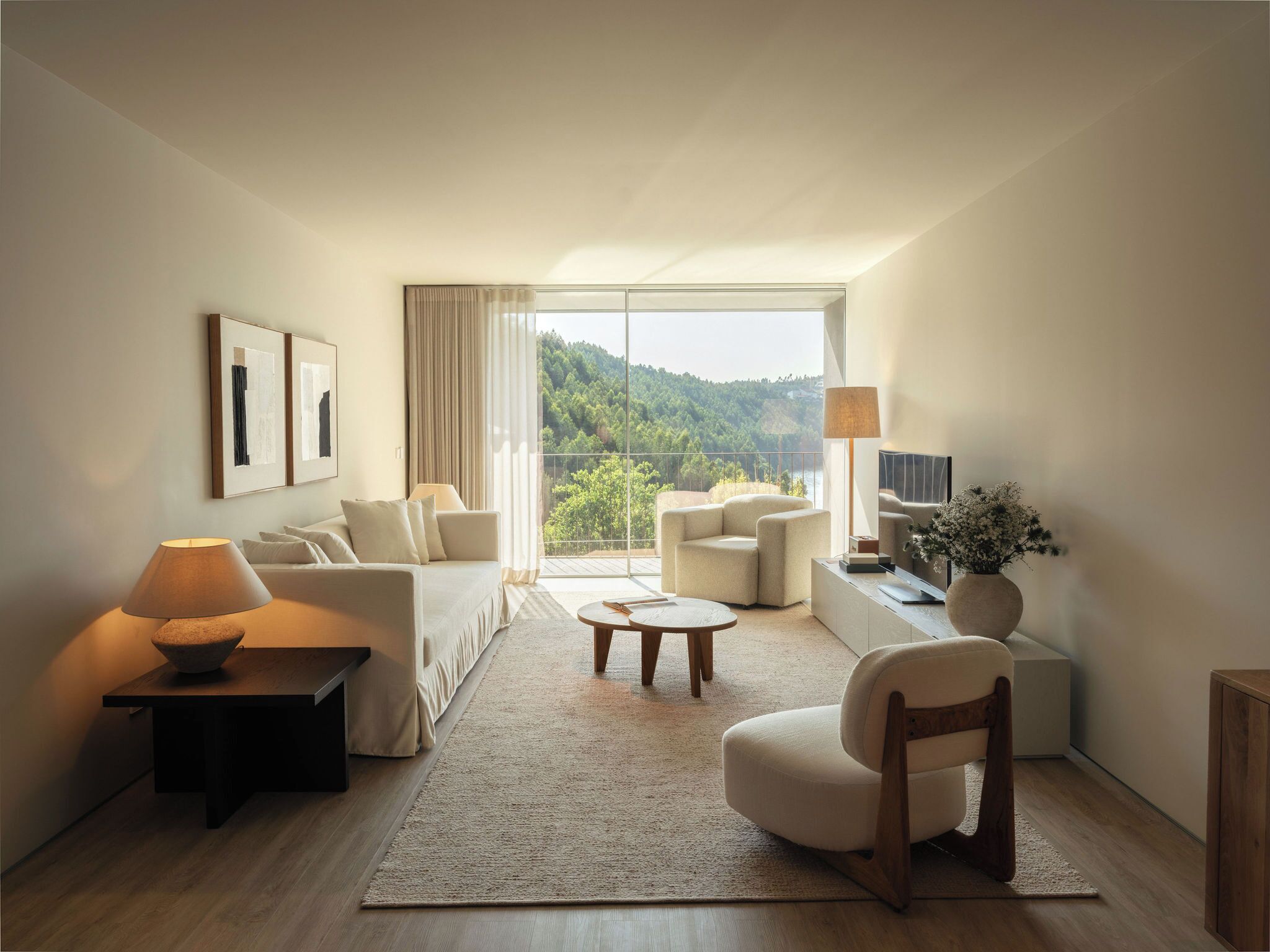 A modern living room with a sofa, armchair, and coffee table, a TV, and a view of a forested area.