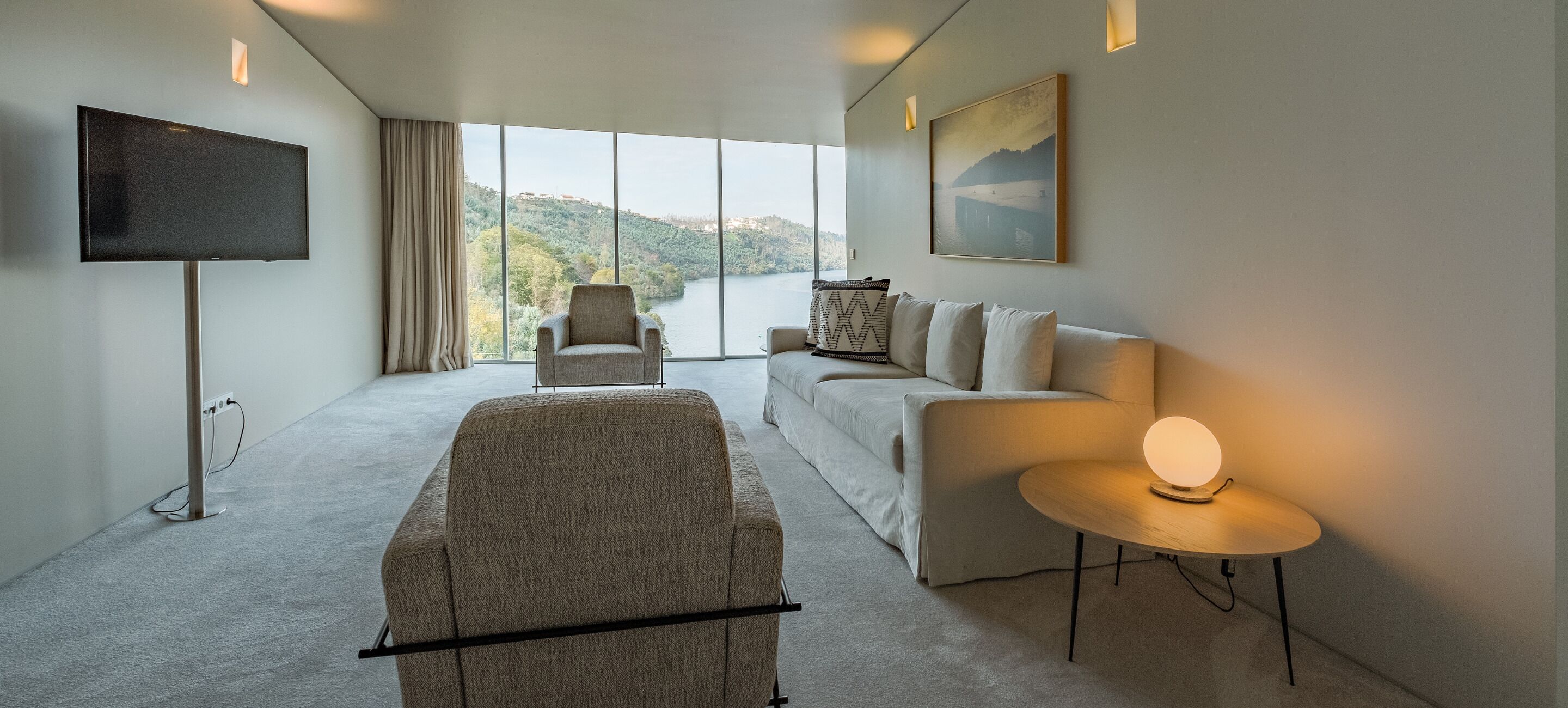 A modern hotel room with a large window, a flat-screen TV, a sofa, a chair, and a small table with a lamp.