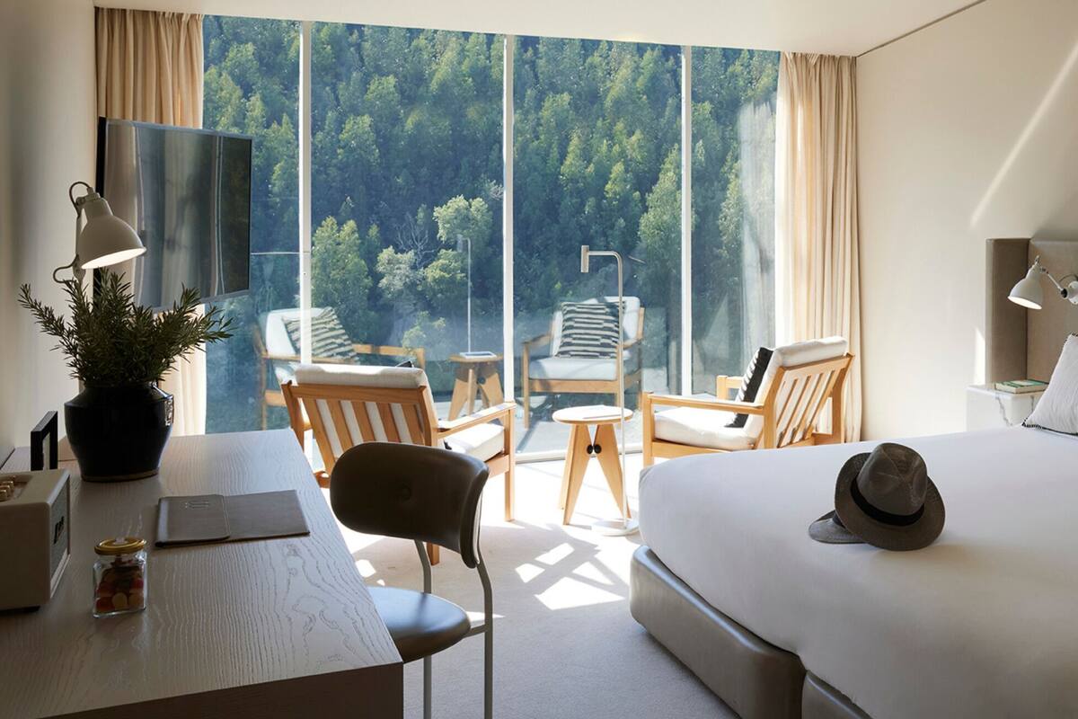 A hotel room with a large bed, a desk with a computer, a chair, and a small table. There is a window with a view of trees and a balcony.