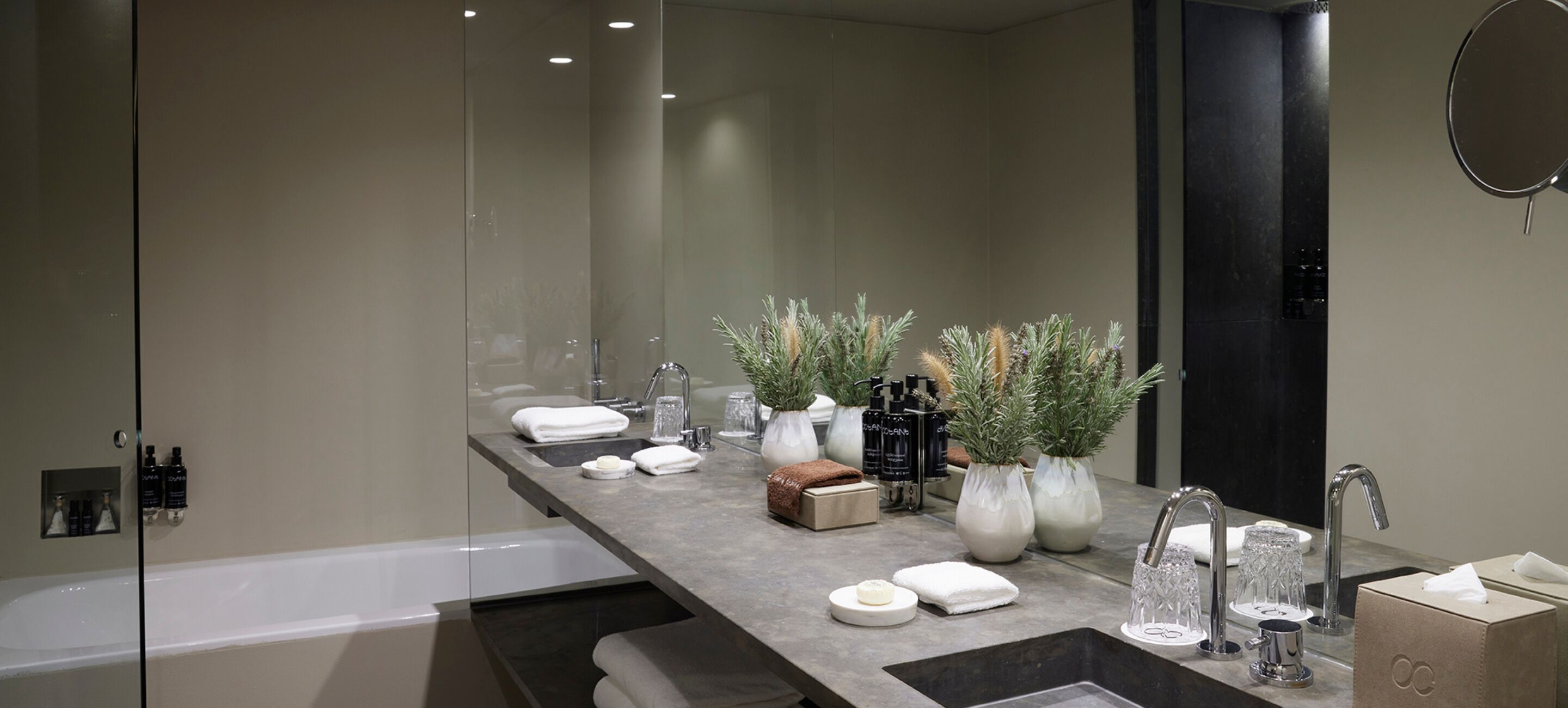 A modern bathroom with a large mirror, a bathtub, and a vanity with toiletries and decorative plants.