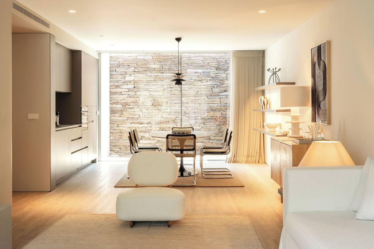 A modern hotel room with a stone wall, a dining area with a table and chairs, a sofa, and a TV.