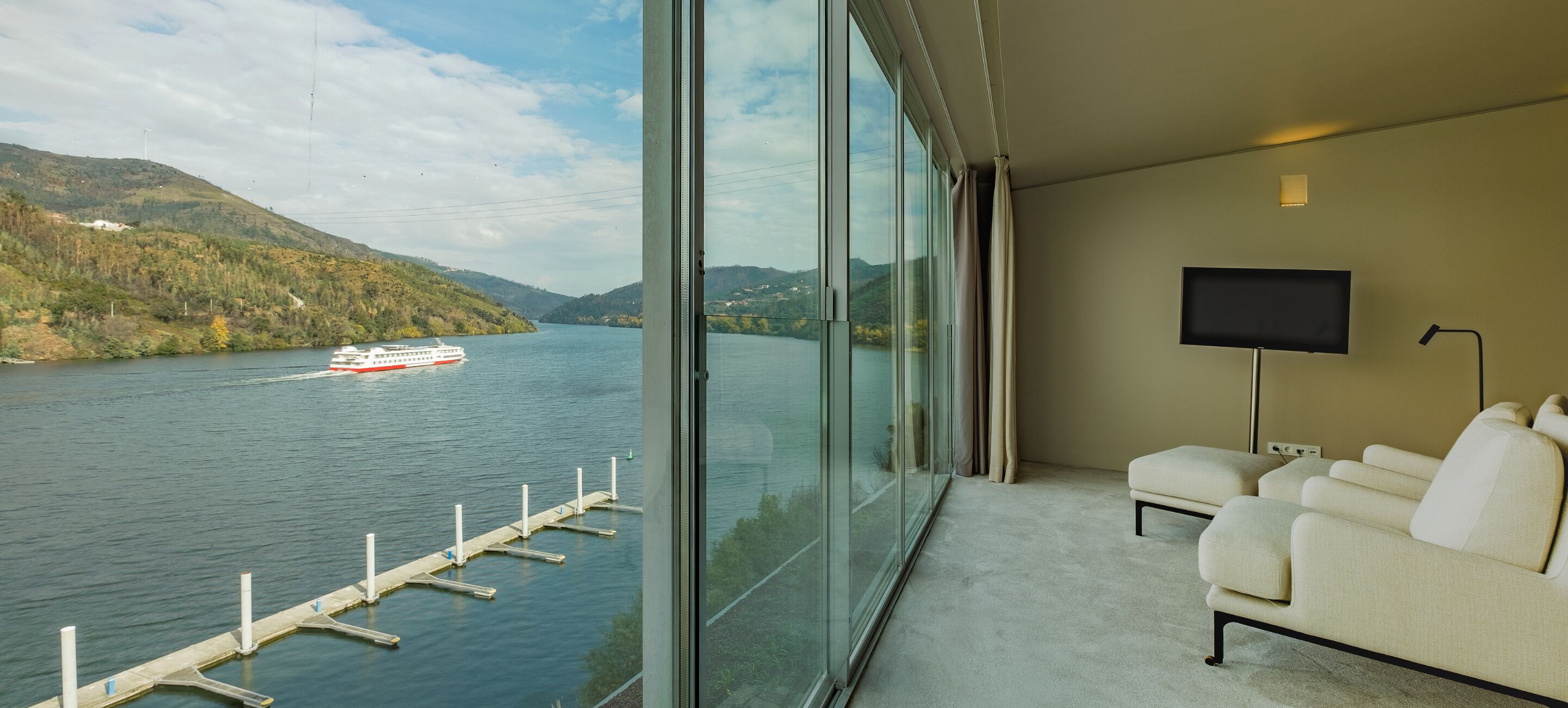 Una habitación de hotel moderna con un ventanal que ofrece vistas a un lago y colinas. Hay un sofá blanco y un televisor de pantalla plana montado en la pared.