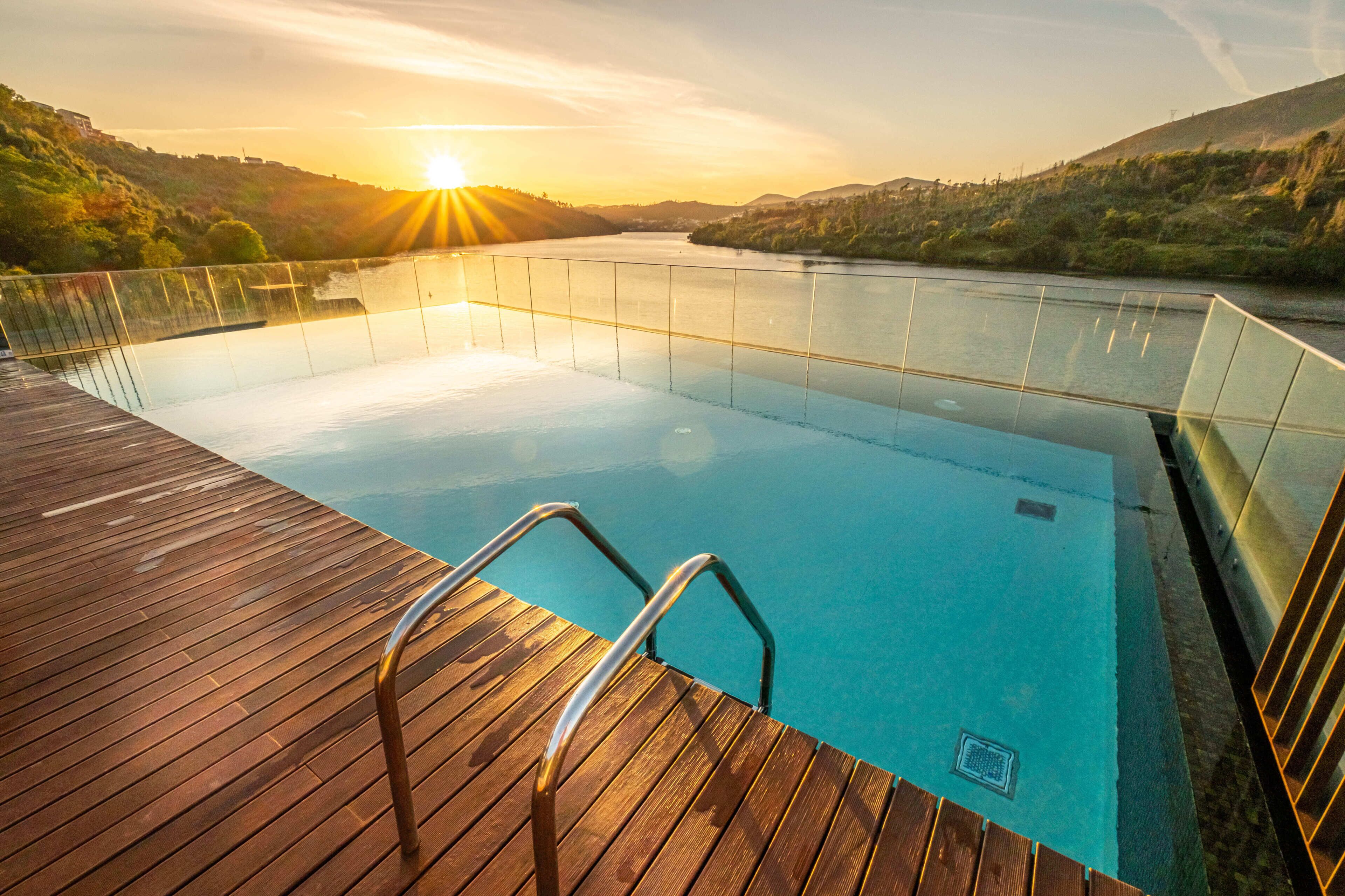 Ein Schwimmbecken mit Holzterrasse und Metallgeländern, umgeben von Hügeln und einem Sonnenuntergang.