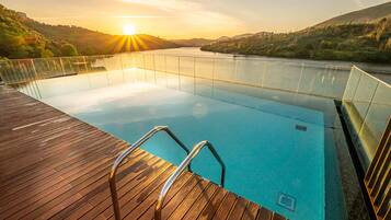 Ein Schwimmbecken mit Holzterrasse und Metallgeländern, umgeben von Hügeln und einem Sonnenuntergang.