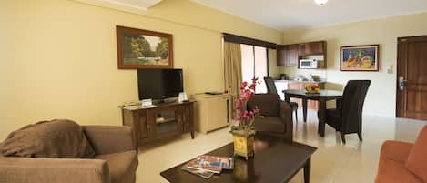 A living room with a sofa, coffee table, and TV. There is a dining area with a table and chairs. The room has a kitchen with cabinets and a microwave.